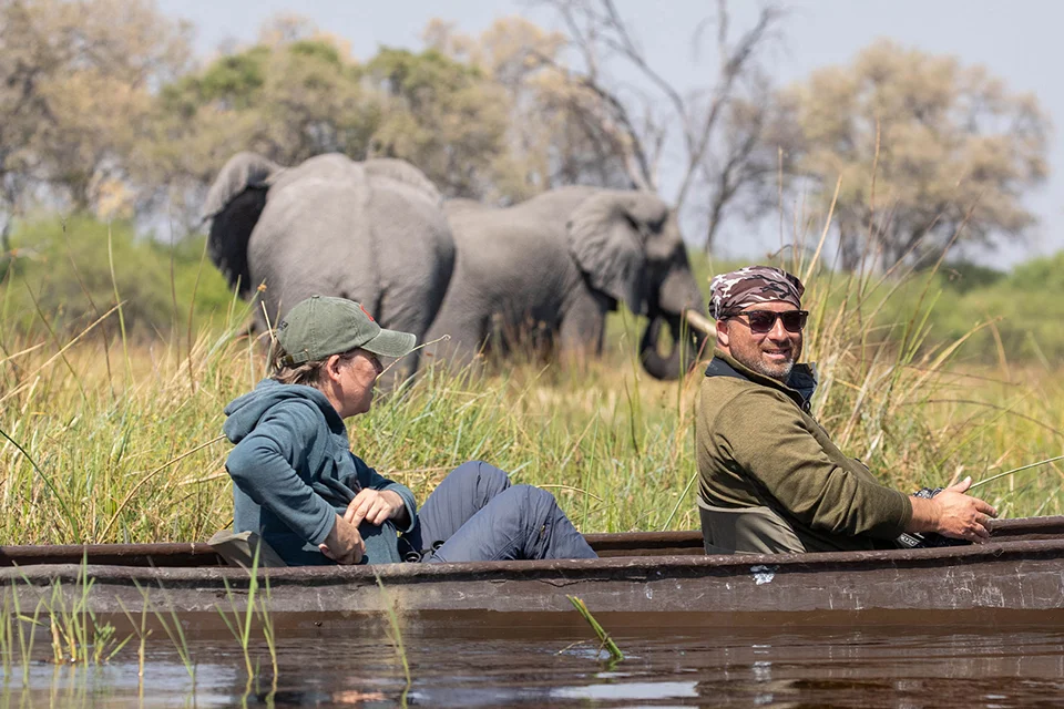 Guests exploring Botswana's Okavango Delta by mokoro as an elephant walks through the surrounding floodplains