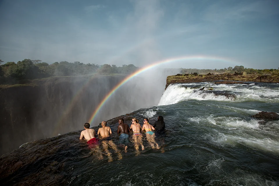 Guests relaxing in Devil's Pool at the edge of Victoria Falls with a rainbow in the background