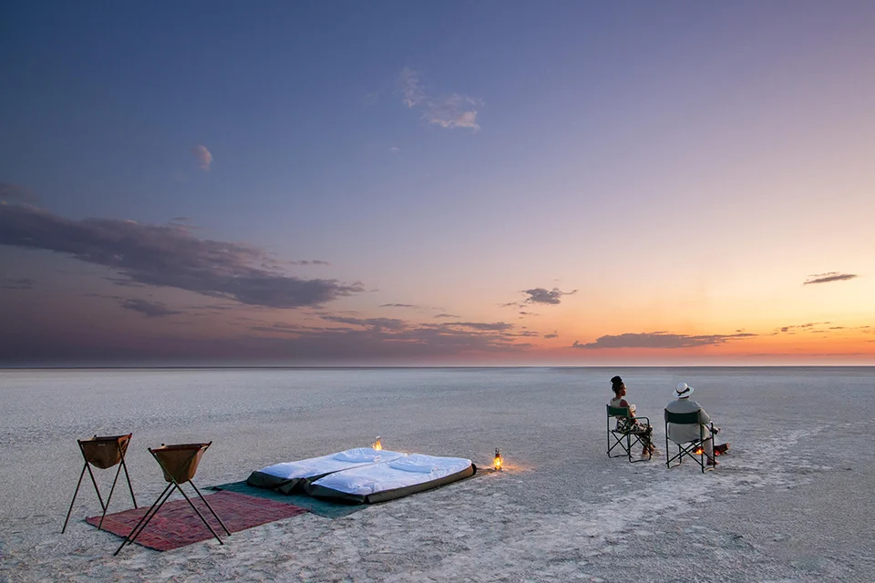 Guests relaxing beside an outdoor sleep-out bed at sunset on Botswana’s Makgadikgadi Salt Pans