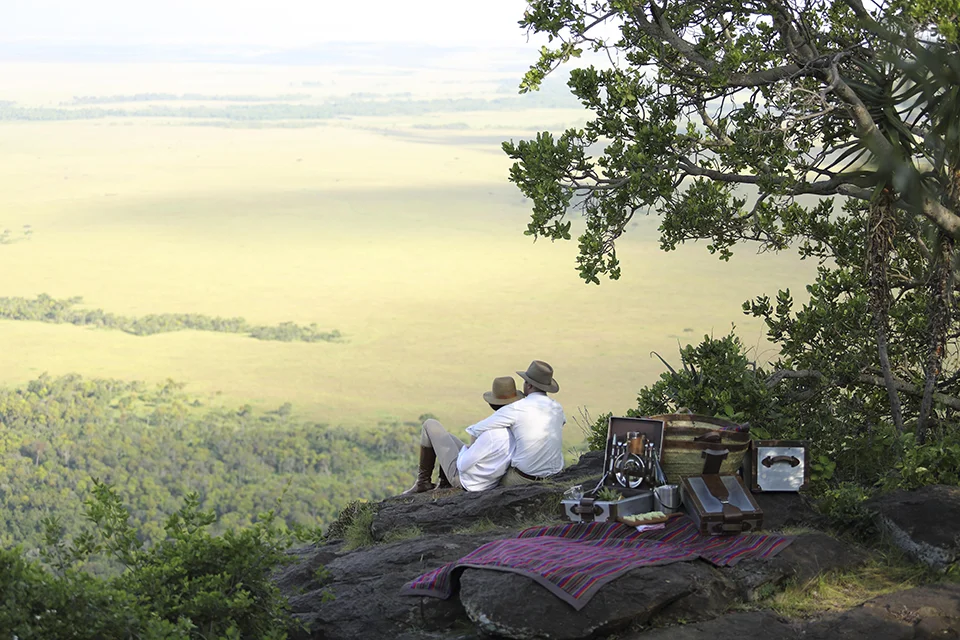 Two guests relax on a rocky viewpoint overlooking the Maasai Mara, enjoying a honeymoon private picnic with sweeping views across the savannah.