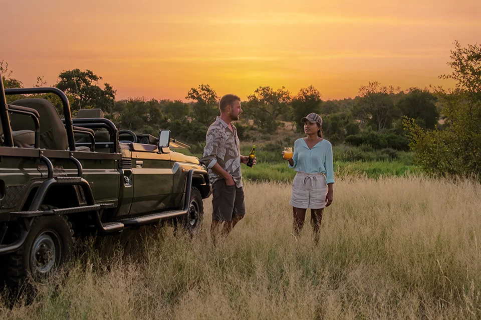 Couple enjoying sunset drinks beside an open safari vehicle in the African bush, a romantic honeymoon safari moment surrounded by golden grasslands and warm evening light