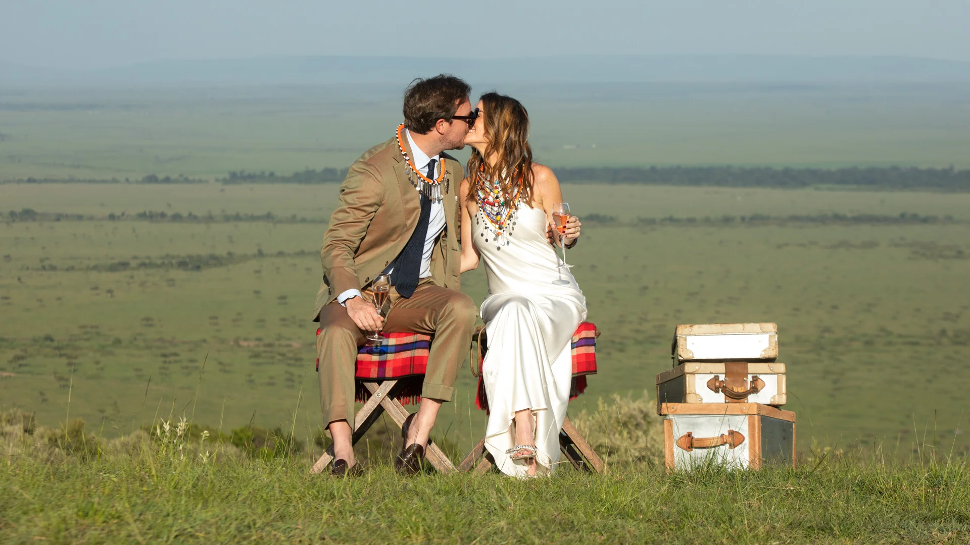 Newlywed couple share a kiss while toasting with champagne on a scenic hilltop overlooking the Maasai Mara, seated beside vintage safari trunks during a romantic safari celebration