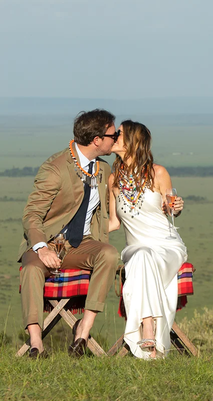 Newlywed couple share a kiss while toasting with champagne on a scenic hilltop overlooking the Maasai Mara, seated beside vintage safari trunks during a romantic safari celebration