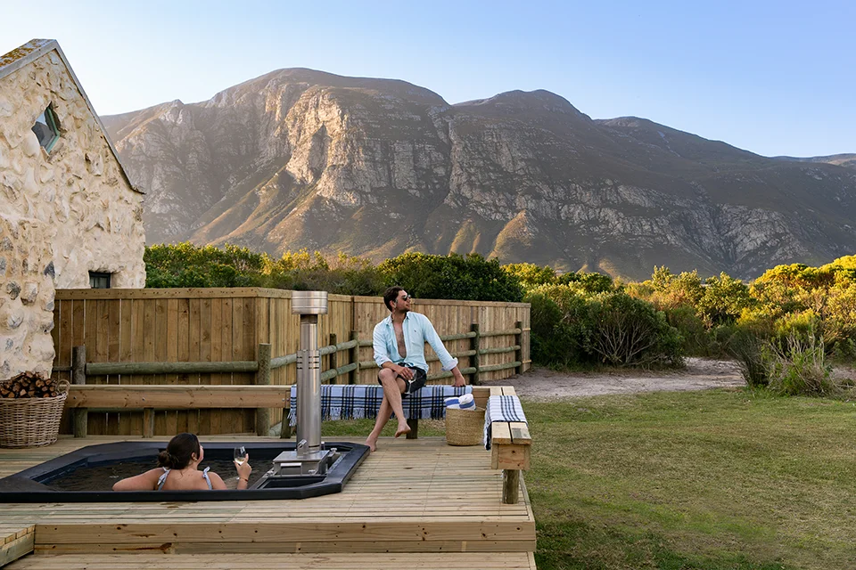 Couple unwind in a private wood-fired hot tub beside a luxury cottage, enjoying mountain views during a romantic South African honeymoon getaway