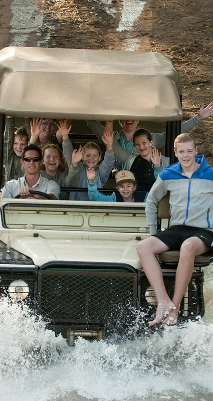 A safari vehicle drives through a shallow water crossing, sending up large splashes as a group of smiling adults and children wave toward the camera
