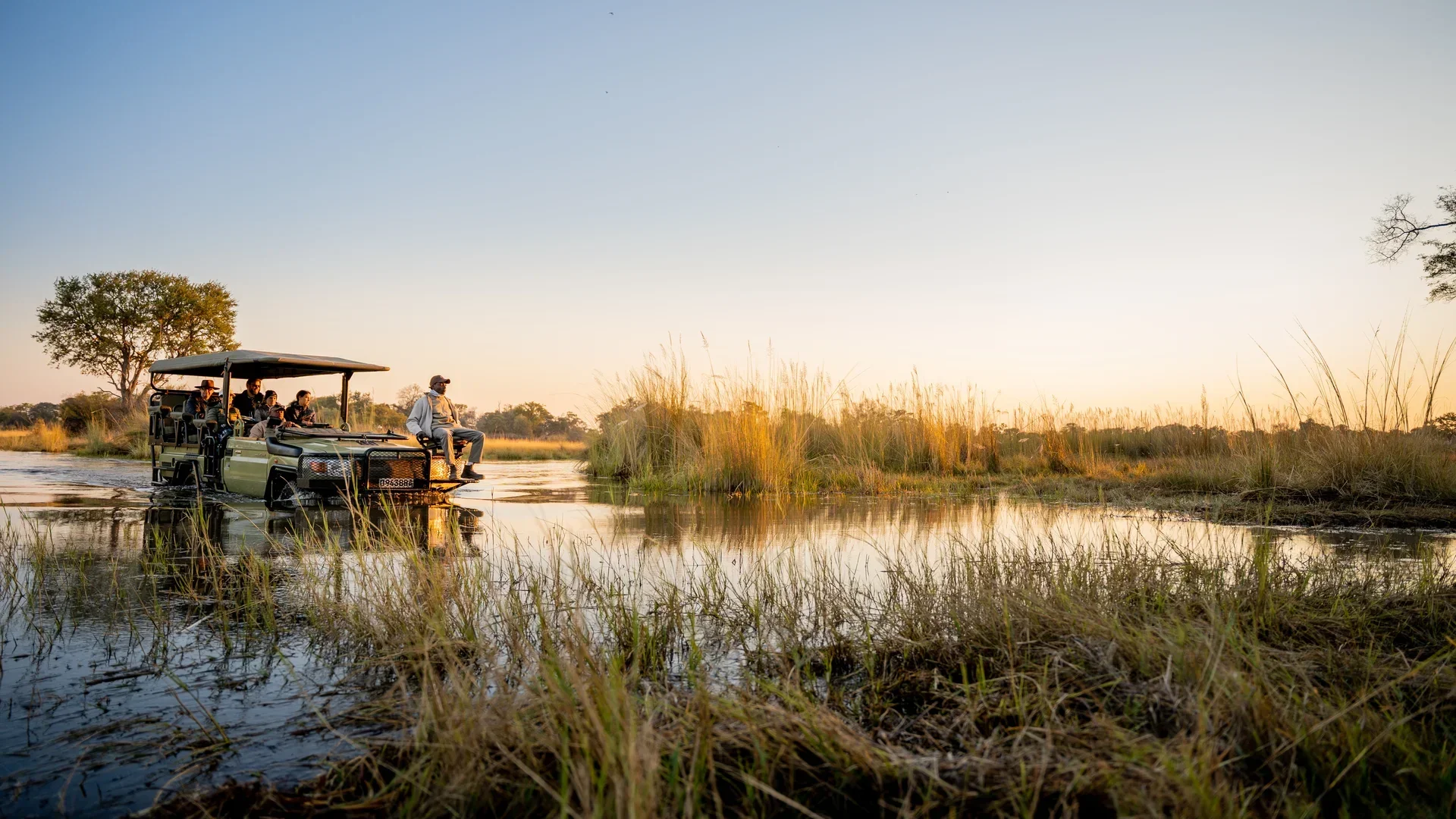 Safari vehicle crossing shallow floodwaters in Botswana's Okavango Delta during a golden sunset game drive