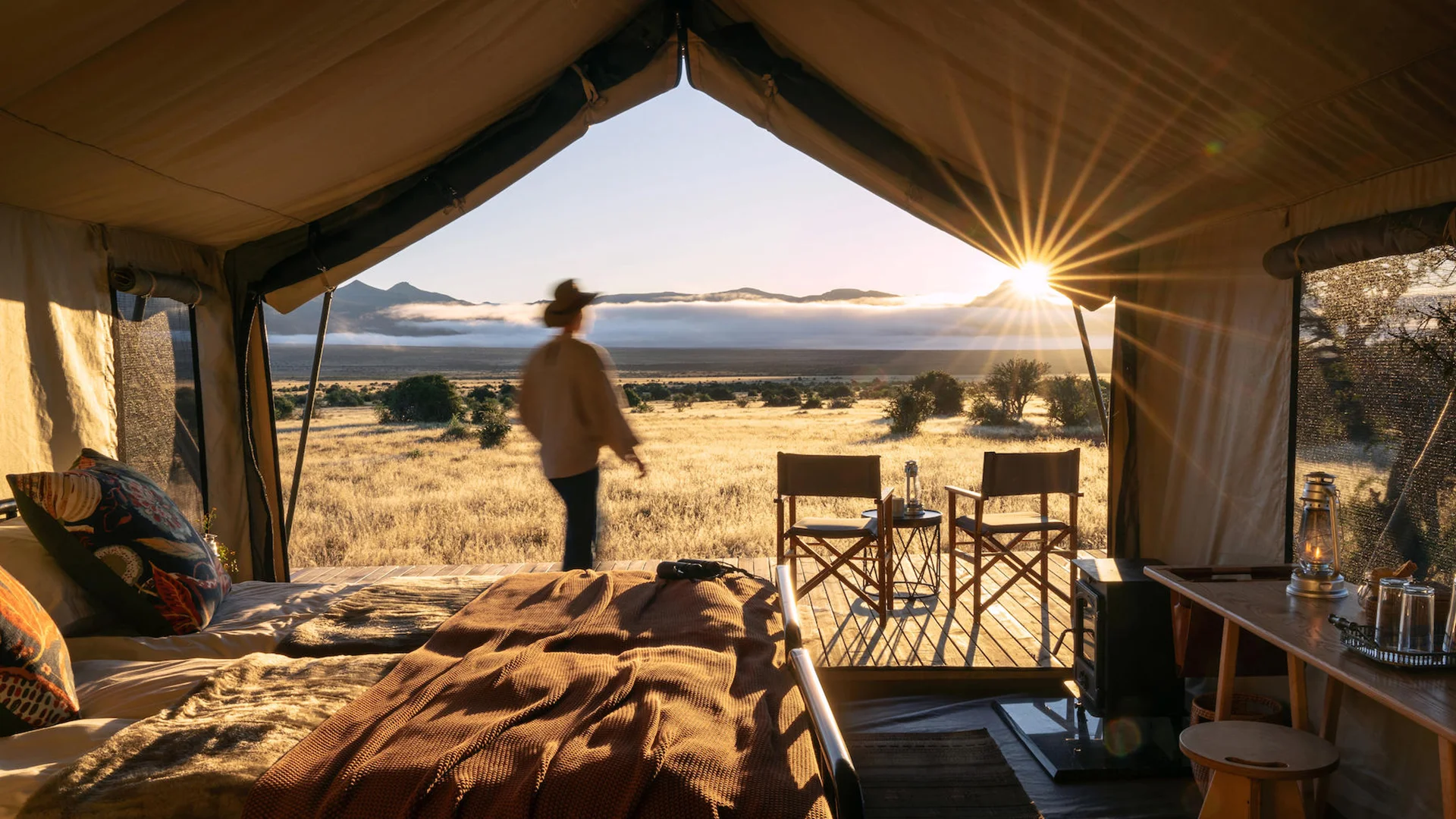 Luxury safari tent overlooking the Samara Karoo plains at sunrise, with a guest stepping onto the private deck and mountain views beyond