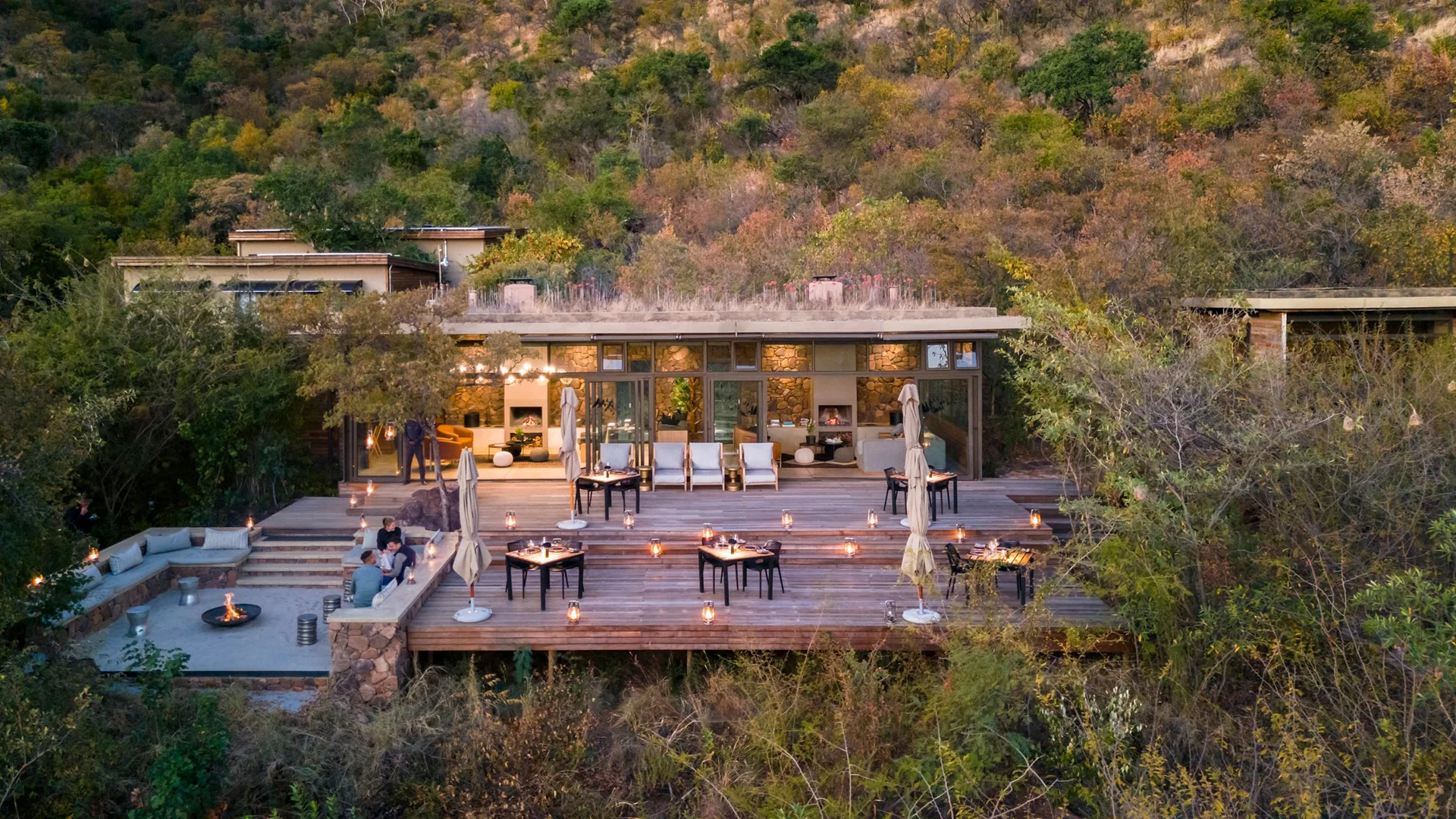 Outdoor dining deck at a safari lodge, surrounded by dense bushveld and set with candlelit tables