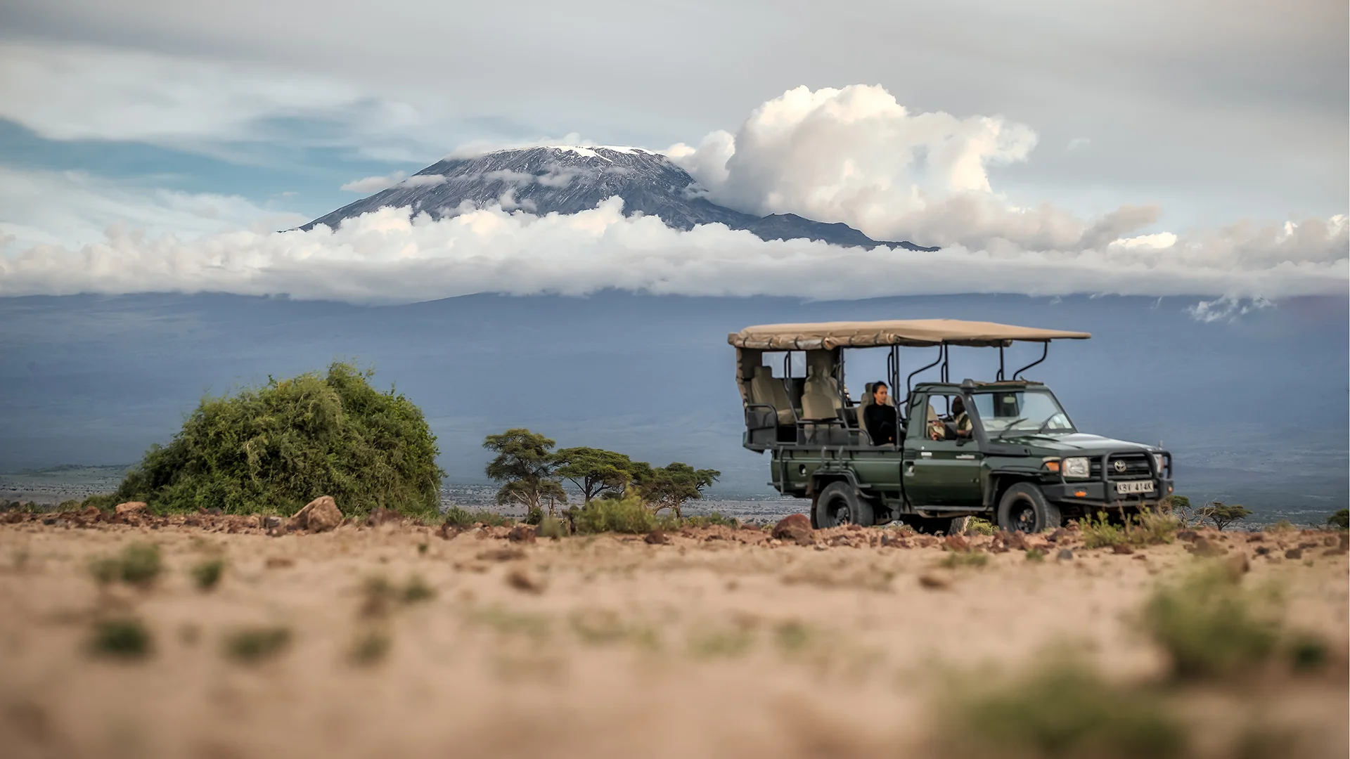 Open-air safari vehicle on a rocky plain with Mount Kilimanjaro rising through low clouds in the background