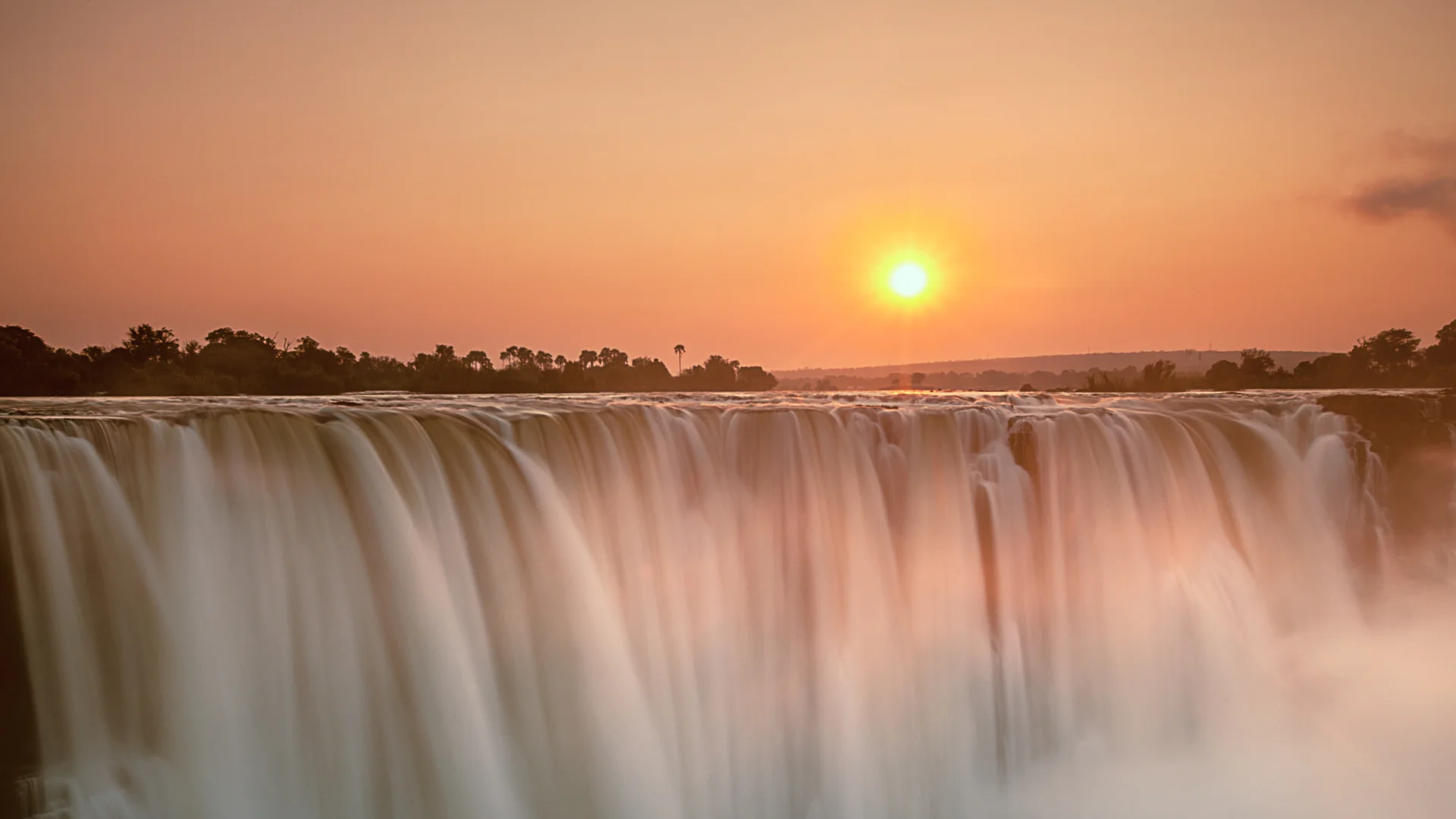 Victoria Falls at sunset, with golden light illuminating the cascading water and rising mist along the Zambezi River